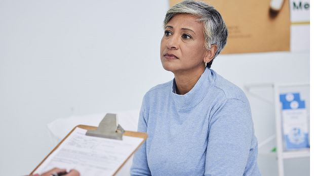 A woman with short hair dressed in a light blue sweater in a doctor's office looking at someone (not seen) taking notes on a clipboard.