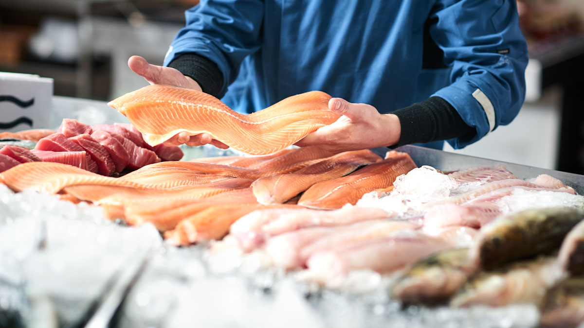 A close up of hands holding a long slice of fresh fish. An assortment of fresh, raw fish are set out on ice at a fish market.