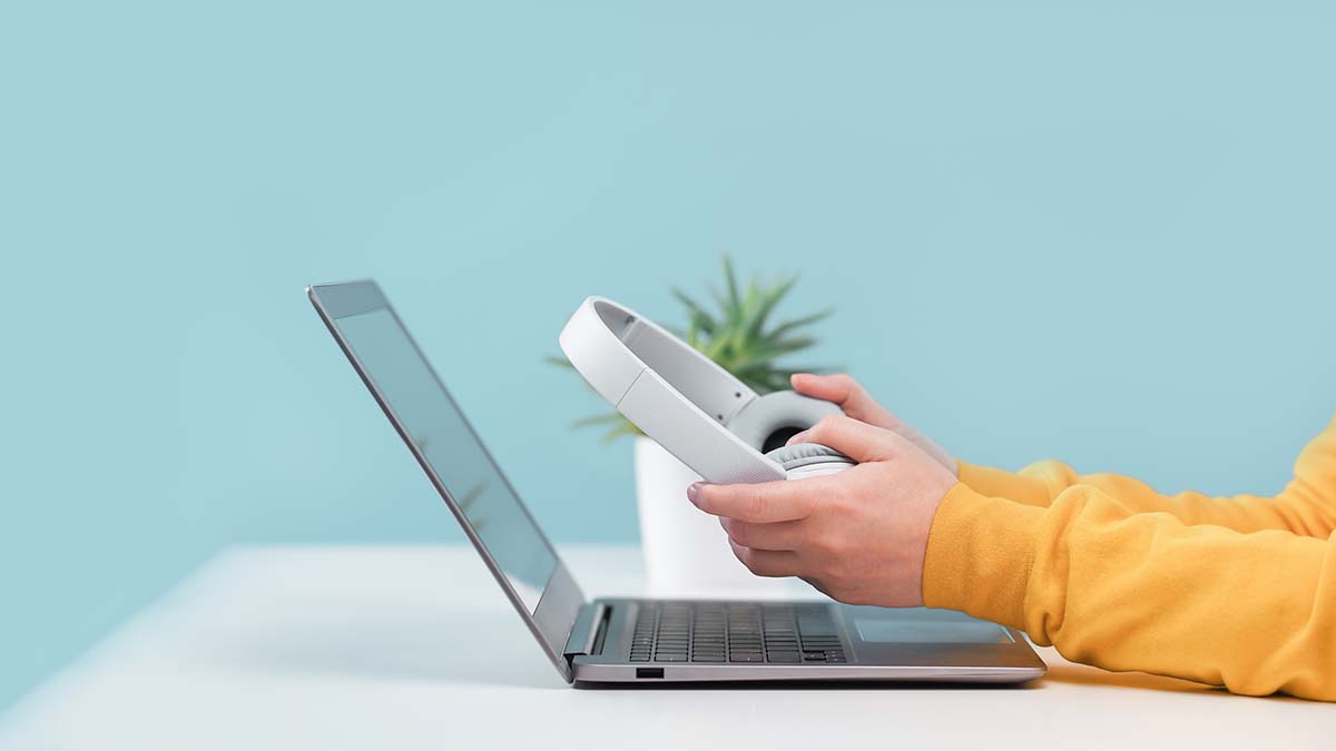 Side view of slim laptop and hands with wireless headphones on grey desk. Blue background. Distant learning. working from home, online courses or support. Audio podcast. vlogger or blogger banner