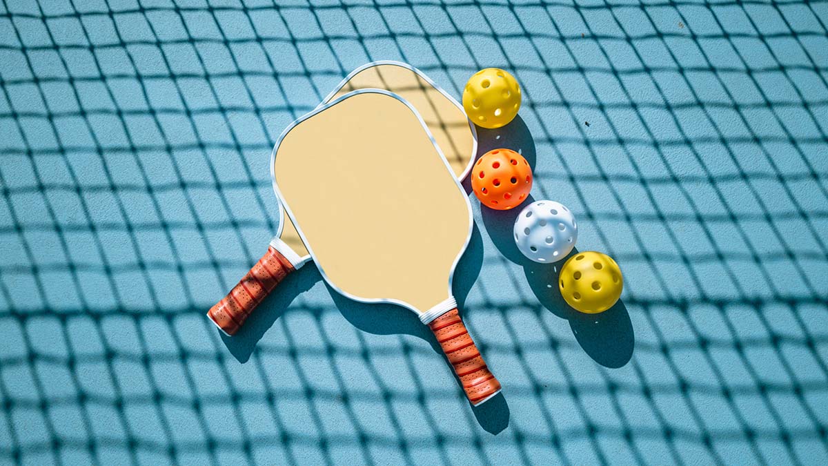 Pickleball paddles and balls laying on the ground with a shadow of the net.