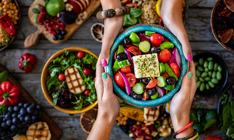 Two women hold a Greek salad, surrounded by healthy foods including fruits and vegetables.